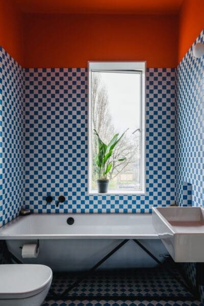 A bathroom featuring walls tiled with a blue and white checkerboard pattern. A white bathtub with black fixtures sits below a window. A white sink is visible to the right of the tub. The ceiling is painted a bright orange. The floor has a black and white geometric pattern. A toilet is visible on the left. A green plant sits on the windowsill.