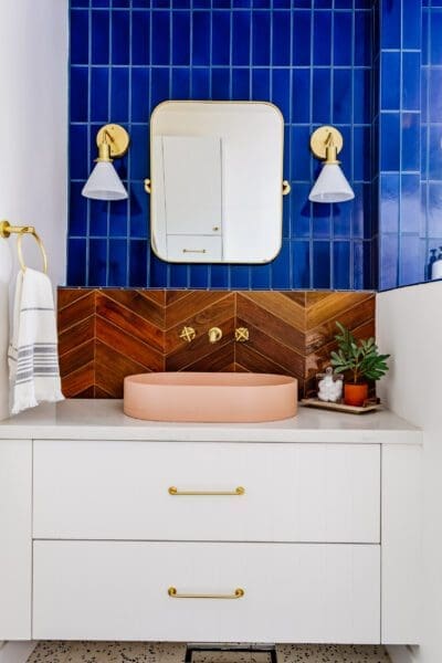 A modern bathroom vanity featuring a white cabinet with gold hardware, a light pink oval sink, and a light-toned countertop. A backsplash of warm-toned wood in a herringbone pattern adds a natural element. Above the sink, a gold-framed rectangular mirror is flanked by two white and gold sconces. The wall behind the mirror is tiled with vertical rows of bright blue subway tiles. A gold towel bar hangs to the left of the mirror, and a small potted plant sits on the counter to the right. The floor appears to have a speckled pattern.