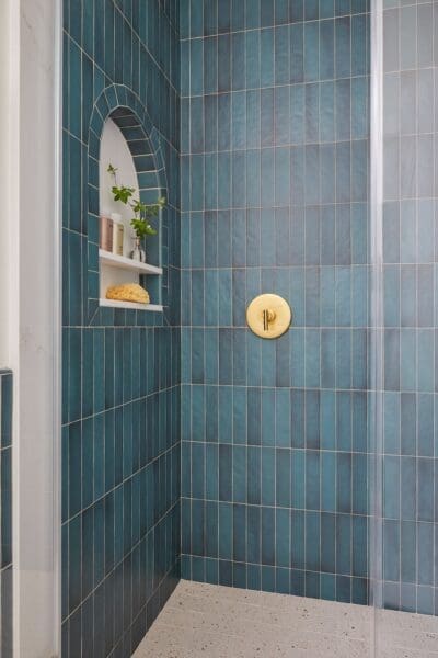 A modern bathroom featuring a shower with vertical teal tiles, a gold showerhead and fixtures, and a built-in shelf holding bottles and a sponge. The floor is tiled with small, colorful geometric tiles, and a white toilet is visible. A light wood vanity with a white countertop and a round mirror is on the left side of the frame.