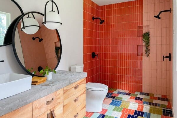 A vibrant and modern bathroom with a playful color scheme. The shower area features two distinct sections: one tiled with vertical rows of glossy orange-red subway tiles and the other with vertical rows of matte light pink subway tiles. Each section has a black showerhead and black fixtures. A built-in niche with a plant is visible in the light pink tiled section. The bathroom floor is tiled with a colorful patchwork of small rectangular tiles in shades of blue, green, orange, pink, and white. A white toilet sits next to the shower. A wooden vanity with a concrete countertop and a rectangular white sink is on the left. A large round mirror with a black frame and two hanging white pendant lights is mounted above the sink. A small potted plant and a folded orange towel are on the countertop. The walls are painted white. The overall design is bold, contemporary, and full of personality.