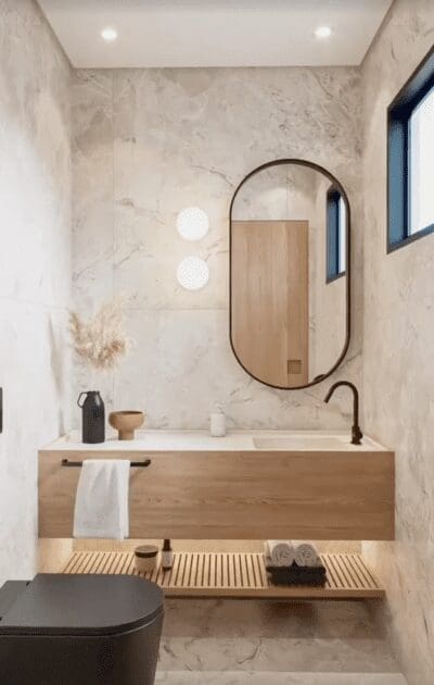 A modern bathroom featuring a light wood vanity with a white countertop and a black, oval-shaped Viva-Black 55 mirror mounted above it. Two round white sconces flank the mirror. A black faucet and a white towel hanging from a black towel bar are visible on the vanity. A dark gray toilet is in the foreground, and a slatted shelf beneath the vanity provides storage. The walls are covered in light gray marble-look tiles. A blue-framed window is visible on the right.