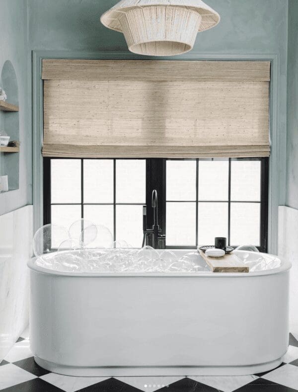 A luxurious bathroom featuring a freestanding white oval bathtub filled with bubbles. A black gooseneck faucet is mounted at one end of the tub. A wooden bath caddy rests across the tub, holding a white soap dish and a black object. A large window with black frames and multiple panes is behind the bathtub, covered by a light tan woven blind. Above the window, a unique white woven pendant light fixture hangs from the ceiling. The walls are painted a soft blue-gray, and the floor is covered in a striking black and white diamond-patterned tile. A glimpse of a built-in arched shelf with white objects is visible on the left wall. The overall atmosphere is serene and spa-like.