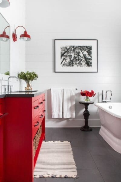 A bathroom featuring a bright red vanity with black hardware and a dark countertop. Two red gooseneck sconces illuminate a mirror above the vanity. White shiplap walls contrast with the dark gray tiled floor. A small black table with red flowers sits next to a white clawfoot bathtub. A striped rug lies on the floor. A black and white framed print hangs on the wall.