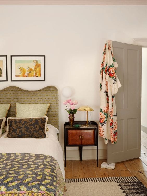 A charming and eclectic bedroom corner featuring a bed with a textured, scalloped headboard in muted green and yellow tones. The bed is dressed with white linens, patterned throw pillows including a dark floral one, and a colorful quilt with yellow and blue leaf-like designs. A small, vintage-style nightstand with a dark wood top and two brown cabinet doors sits beside the bed. On the nightstand, there is a small brass lamp with a pleated shade, a vase of pink tulips, and a small white object. A patterned rug in cream and black lies partially under the bed on a wooden floor. A light gray door is slightly ajar, revealing a glimpse into another room. A floral patterned robe hangs on the back of the door. Above the bed, two framed pieces of artwork with playful animal illustrations adorn the white wall. A small, round wall-mounted light fixture is also visible above the nightstand.