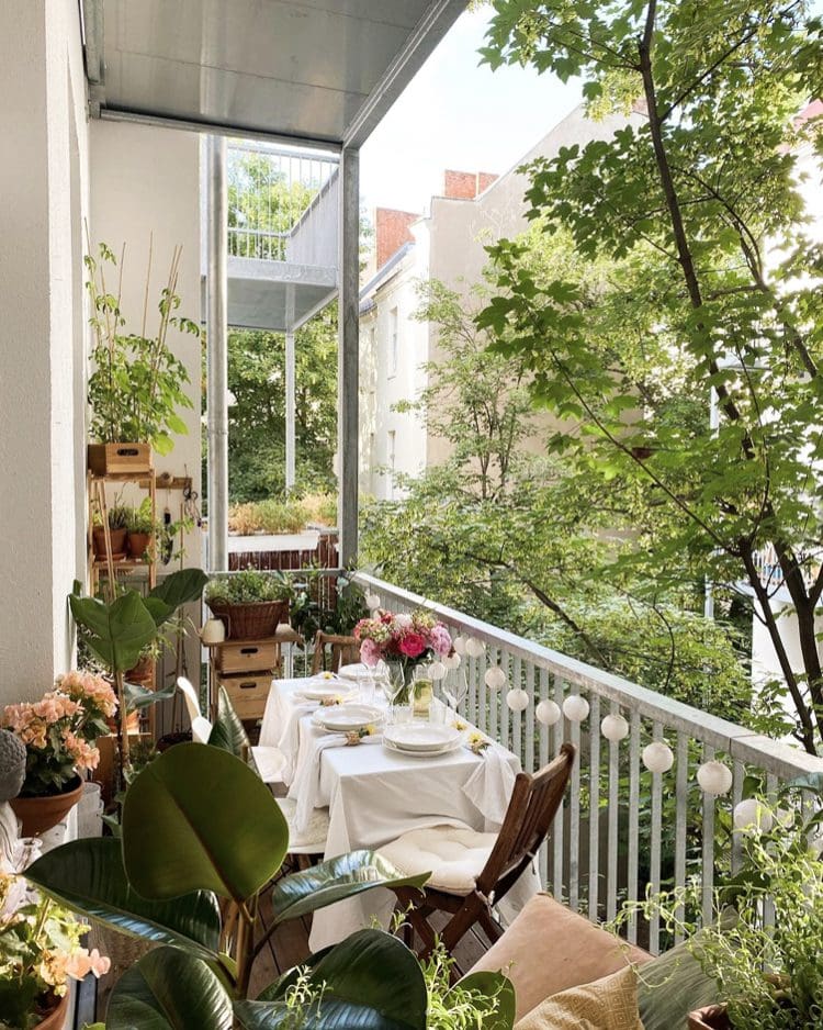 A charming and plant-filled balcony in Valparaiso, Indiana, set for an intimate outdoor meal. A small rectangular table is covered with a white tablecloth and set with white plates, silverware, and simple white napkins. A glass vase filled with pink and white peonies and other flowers serves as a centerpiece. Two wooden folding chairs with white cushions are placed at the table. The balcony railing is lined with string lights featuring round white bulbs and various potted plants, creating a lush and garden-like atmosphere. Tall leafy trees and glimpses of neighboring buildings are visible beyond the balcony, suggesting an urban setting with a touch of nature. The foreground is filled with large potted plants with vibrant green leaves and delicate peach-colored flowers. A small wooden shelf unit holds additional potted plants. The overall ambiance is bright, airy, and inviting, perfect for enjoying a meal outdoors in a small urban space in Valparaiso. The abundance of plants suggests a love for gardening and creating a natural oasis.