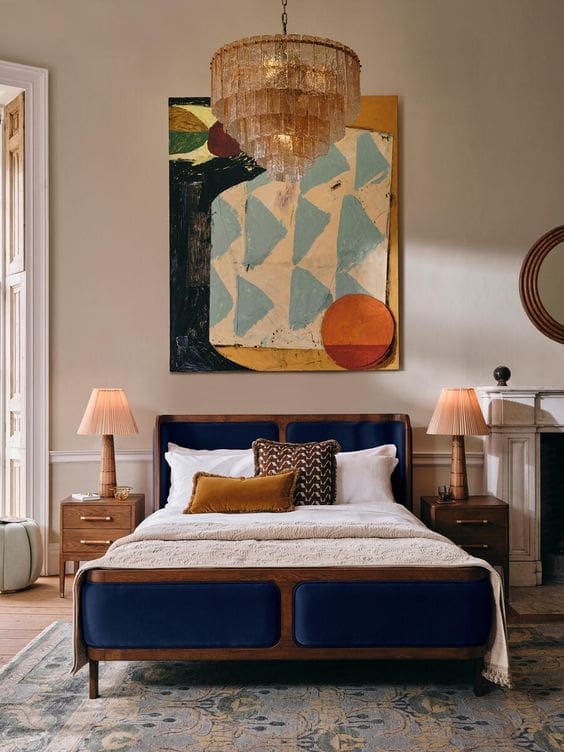 A stylishly decorated bedroom featuring a Soho Home Belsa bed with a deep blue velvet headboard and footboard, and a wooden frame. The bed is dressed with white linens, a textured cream-colored throw, and several decorative pillows in brown and patterned fabrics. Two wooden nightstands with brass accents flank the bed, each topped with a pleated peach-toned lamp. Above the bed hangs a large, multi-tiered chandelier with a textured glass or shell-like appearance. A large abstract painting with muted tones of beige, blue, black, and orange hangs on the wall above the headboard. To the right, a white fireplace mantel with a dark opening and a round mirror above it is visible. The room has light beige walls and a patterned rug in shades of blue, beige, and brown covers the wooden floor. Natural light enters from a window on the left, partially obscured by a light-colored curtain. The overall aesthetic is luxurious and contemporary with a touch of vintage charm