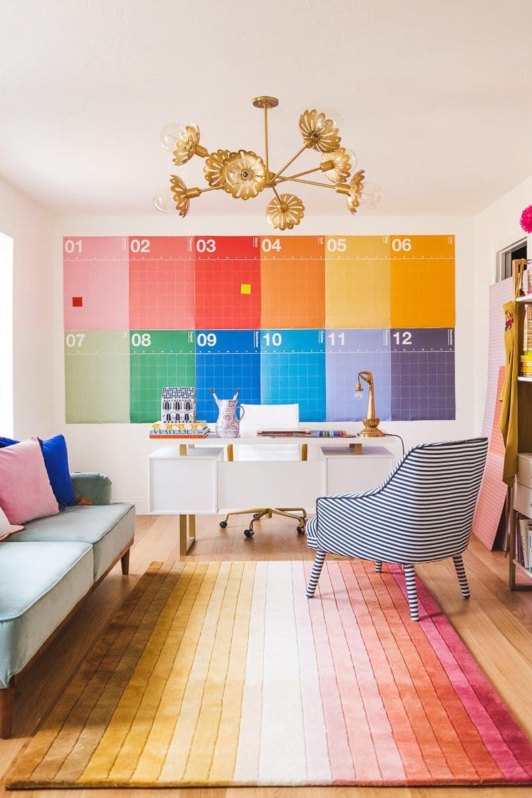 A bright and colorful home office with a large wall calendar featuring a spectrum of colors. A white desk with gold accents sits in front of the calendar, and a striped chair adds a playful touch.