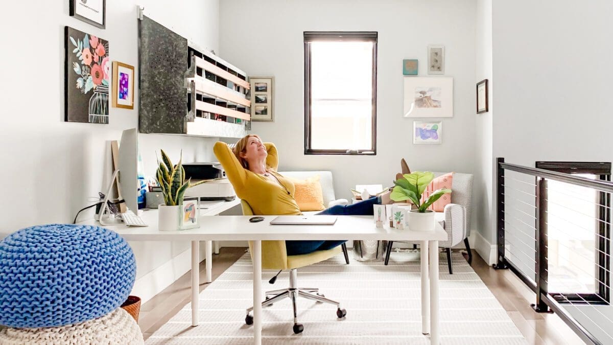 A bright and airy home office featuring a woman relaxing in a yellow chair. She leans back with her hands behind her head, looking up towards a window. A white desk stretches across the frame, holding a laptop, a computer monitor, and potted plants. A blue knitted pouf and a patterned rug add pops of color to the room. Artwork adorns the walls, and a black metal railing separates the office space from another area. The overall feel is modern, creative, and relaxed.