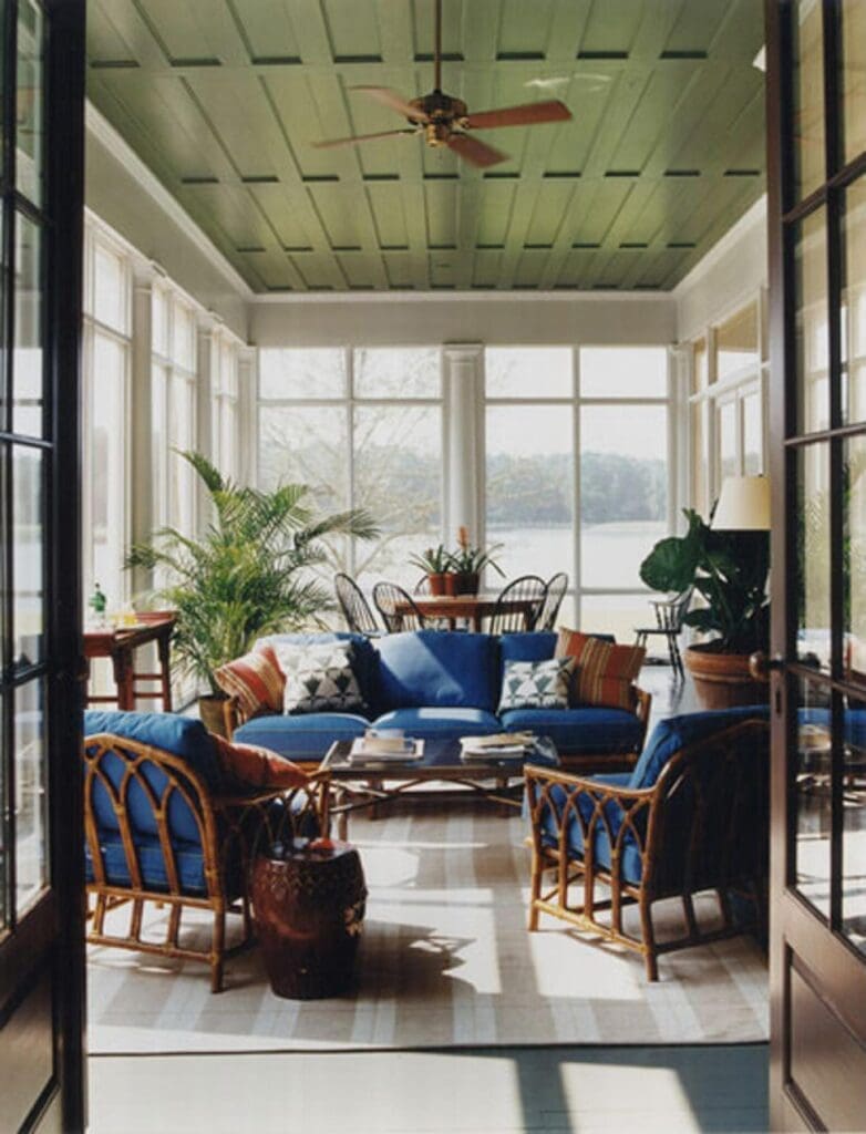A bright and airy sunroom featuring rattan furniture with blue cushions arranged around a light-colored coffee table. The room is enclosed by large windows offering a view of a body of water and trees. A green paneled ceiling with a brown ceiling fan adds a touch of color. Several potted plants bring life to the space, and a striped rug covers the floor. Sunlight streams through the windows, creating shadows and highlighting the textures of the furniture and rug.