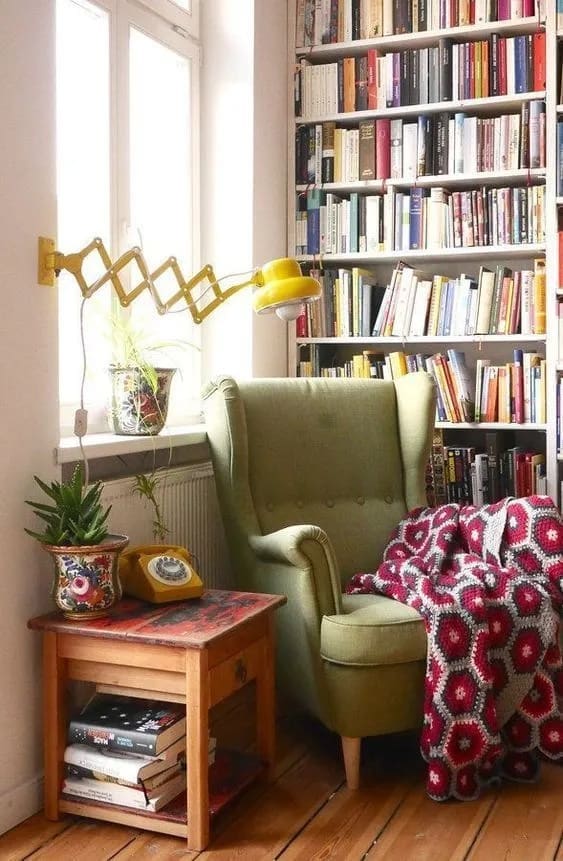 A cozy interior scene featuring a green armchair, a bookshelf filled with books, and a yellow articulated lamp. A small wooden table with books and a houseplant sits next to the chair.