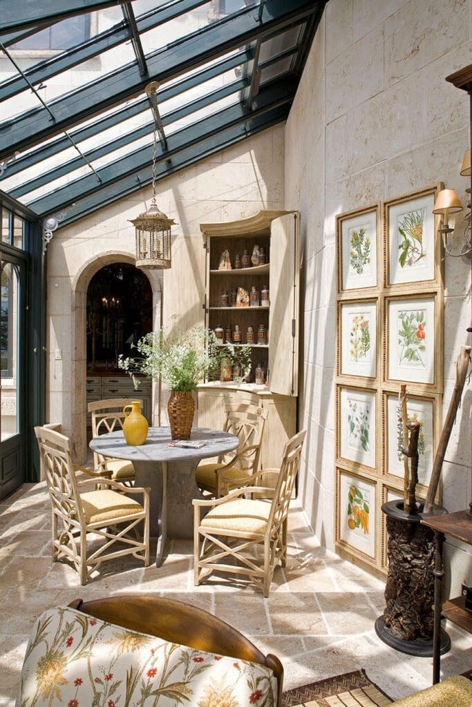 A bright and airy sunroom featuring a round table with chairs, a built-in cabinet, and framed artwork. Sunlight streams through the glass ceiling and windows, illuminating the stone walls and tiled floor. A hanging lantern adds a decorative touch.