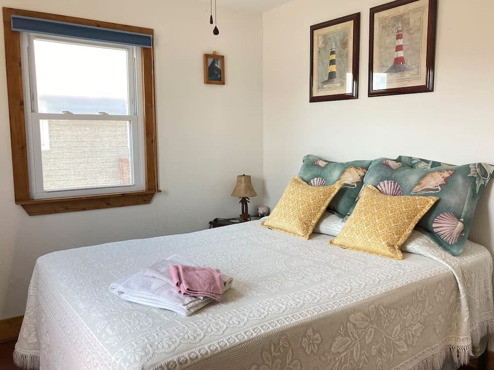 Outdated guest bedroom which could use a design overhaul, with seashell-themed pillows, lighthouse prints, and rustic trim—awaiting a vibrant and modern refresh.
