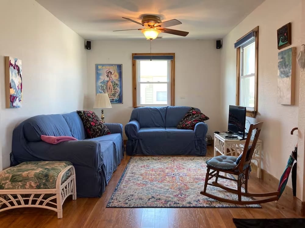 Before image of a dated living room in a beachy home, featuring blue slipcovered sofas, traditional furnishings, and minimal decor—ready for a bold and colorful transformation.