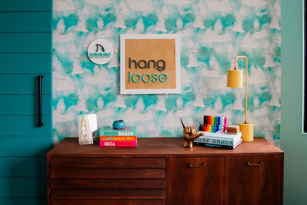 Colorful, beach-themed interior with a wood sideboard against a teal and white abstract wave-patterned wallpaper. Above the sideboard are two wall decorations: one features a shark fin with waves, and the other is a framed sign reading "hang loose." On the sideboard are stacked books with vibrant spines, a photo frame, a blue dish, a golden hand sculpture making the "shaka" sign, a set of rainbow-colored markers, and a yellow desk lamp. A teal sliding door is partially visible on the left.
