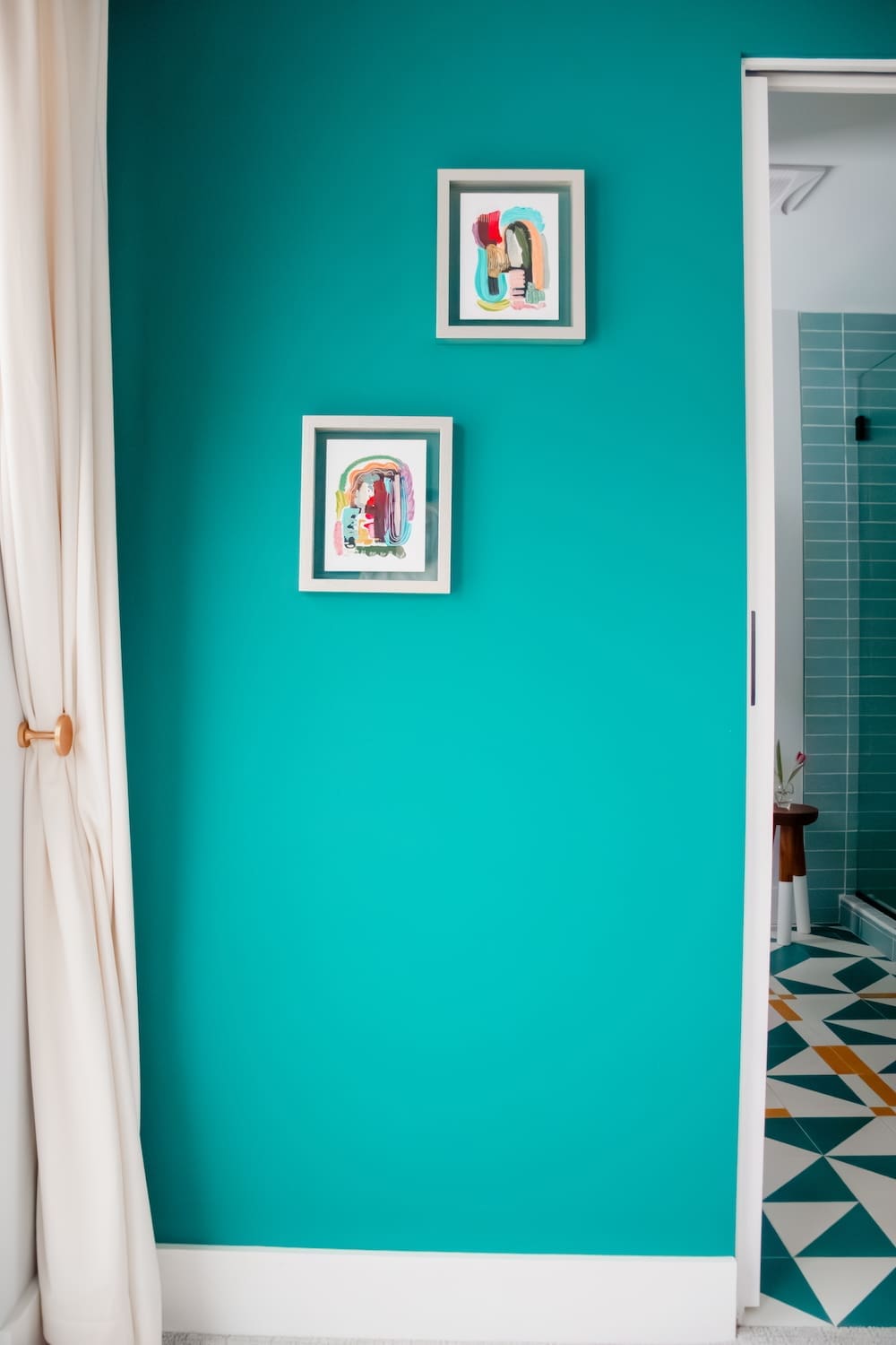 A bright teal bedroom wall with two colorful framed abstract artworks and a peek into an adjoining bathroom with geometric tile flooring, showcasing bold color in timeless home design.
