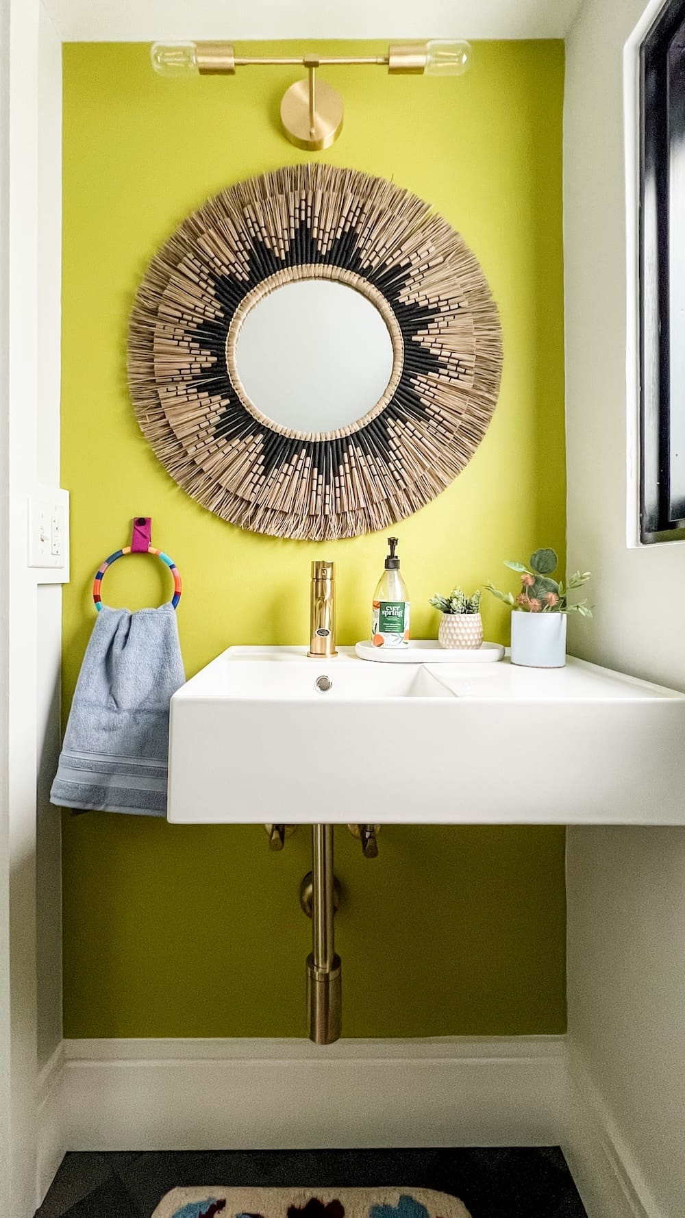 A modern powder room with a bright chartreuse wall, rattan sunburst mirror, brass fixtures, and colorful towel holder, illustrating bold color used in timeless home design.